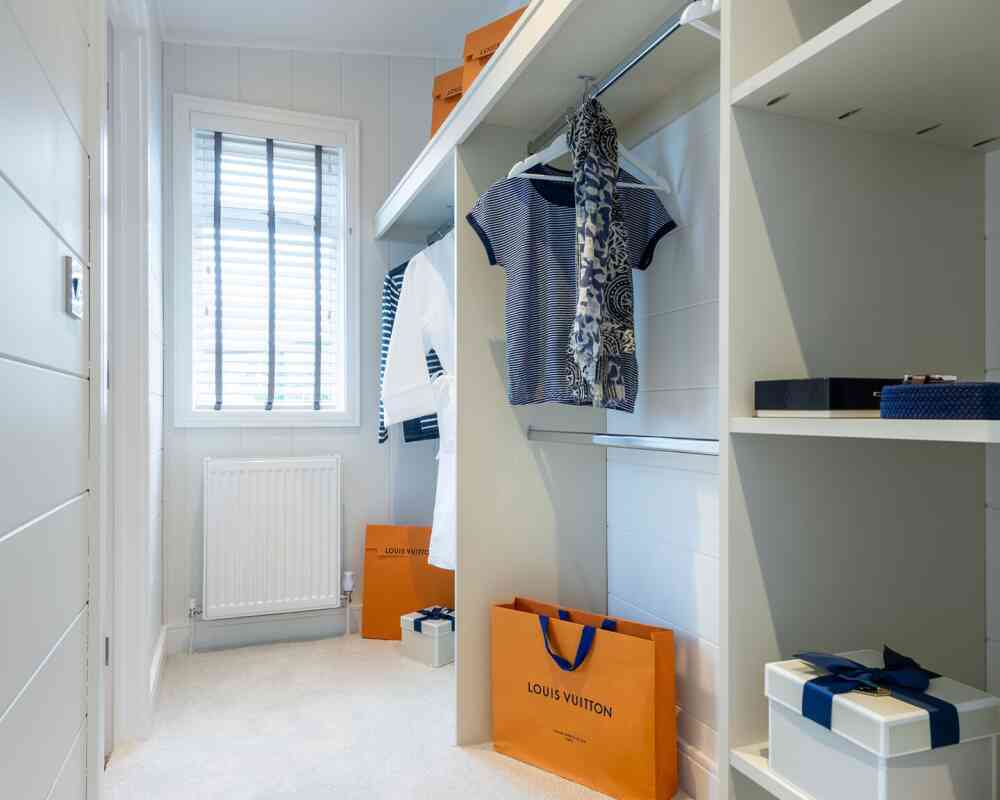 A well-organized walk-in closet featuring hanging clothes, including striped and floral patterns. The space includes shelves with neatly arranged boxes and shopping bags, notably one from Louis Vuitton. A window provides natural light, illuminating the soft carpeted floor.