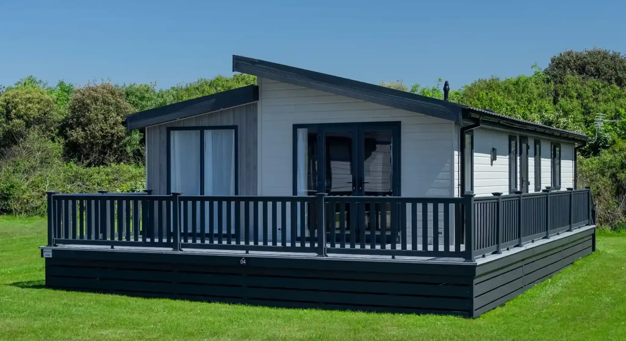 Modern white lodge holiday home with a dark wrap-around deck and grey pitched roof set on a manicured lawn.