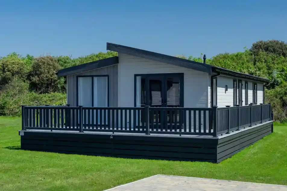 Modern white lodge holiday home with a dark wrap-around deck and grey pitched roof set on a manicured lawn.