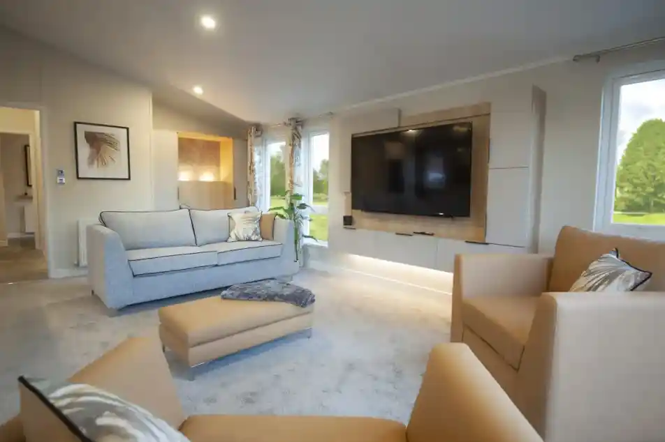 Modern living room with a large wall-mounted TV, a beige armchair, a matching ottoman, a light grey sofa with decorative pillows, and floor-to-ceiling windows allowing natural light inside. The room has neutral-toned walls, carpet flooring, recessed lighting, and a framed wall art piece.