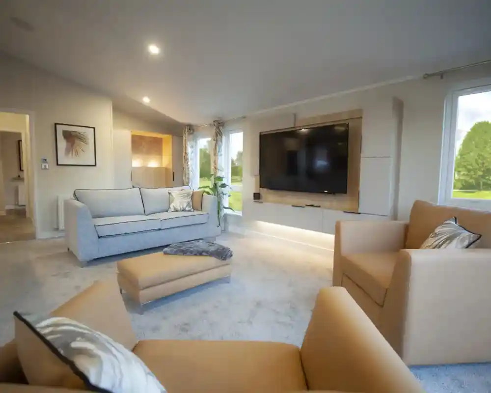 Modern living room with a large wall-mounted TV, a beige armchair, a matching ottoman, a light grey sofa with decorative pillows, and floor-to-ceiling windows allowing natural light inside. The room has neutral-toned walls, carpet flooring, recessed lighting, and a framed wall art piece.
