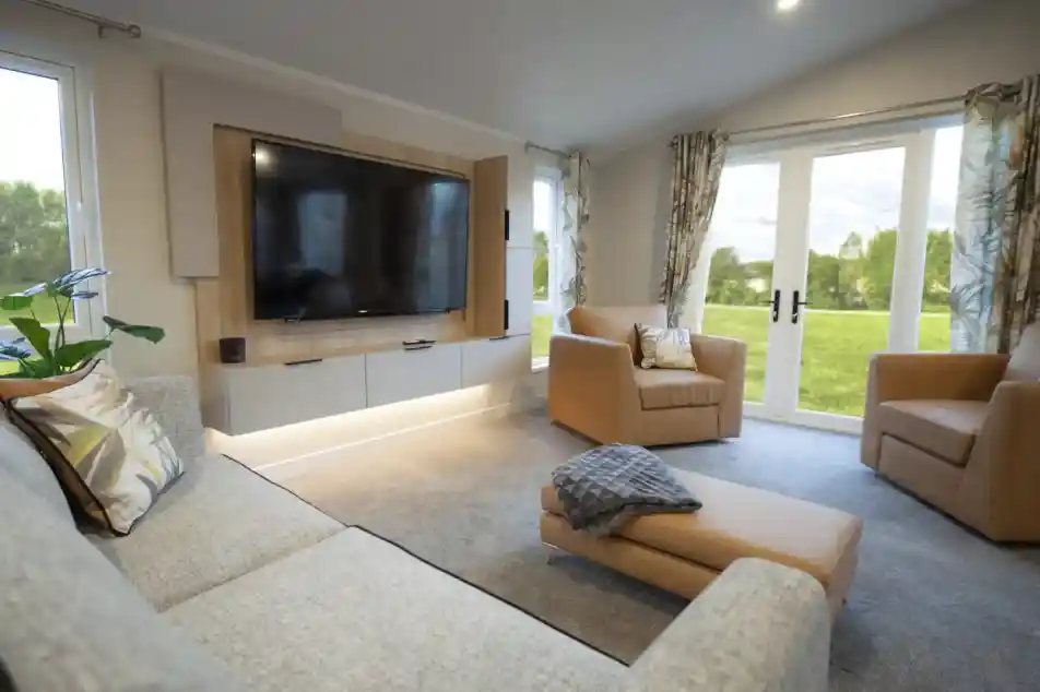 Bright and modern lounge featuring a wall-mounted flat-screen TV with wood panelling, a grey fabric sofa with patterned cushions, two tan armchairs, and a matching ottoman with a throw. Large windows and glass doors with patterned curtains provide views of a green outdoor lawn, filling the room with natural light.