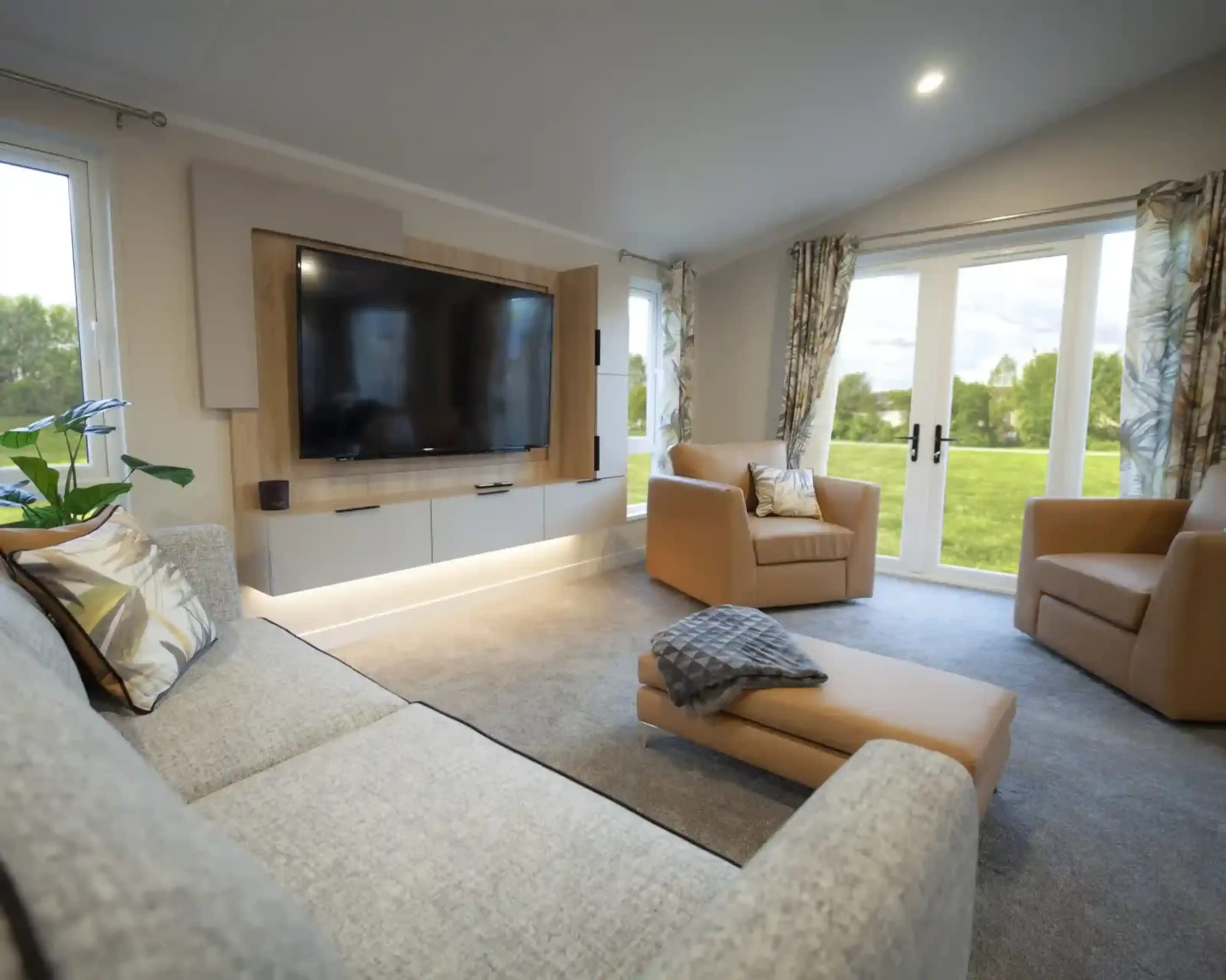 Bright and modern lounge featuring a wall-mounted flat-screen TV with wood panelling, a grey fabric sofa with patterned cushions, two tan armchairs, and a matching ottoman with a throw. Large windows and glass doors with patterned curtains provide views of a green outdoor lawn, filling the room with natural light.