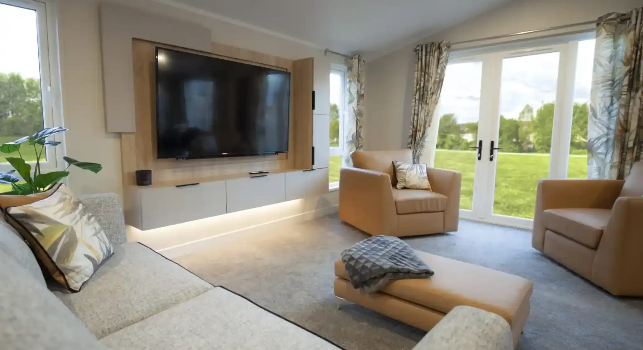Bright and modern lounge featuring a wall-mounted flat-screen TV with wood panelling, a grey fabric sofa with patterned cushions, two tan armchairs, and a matching ottoman with a throw. Large windows and glass doors with patterned curtains provide views of a green outdoor lawn, filling the room with natural light.