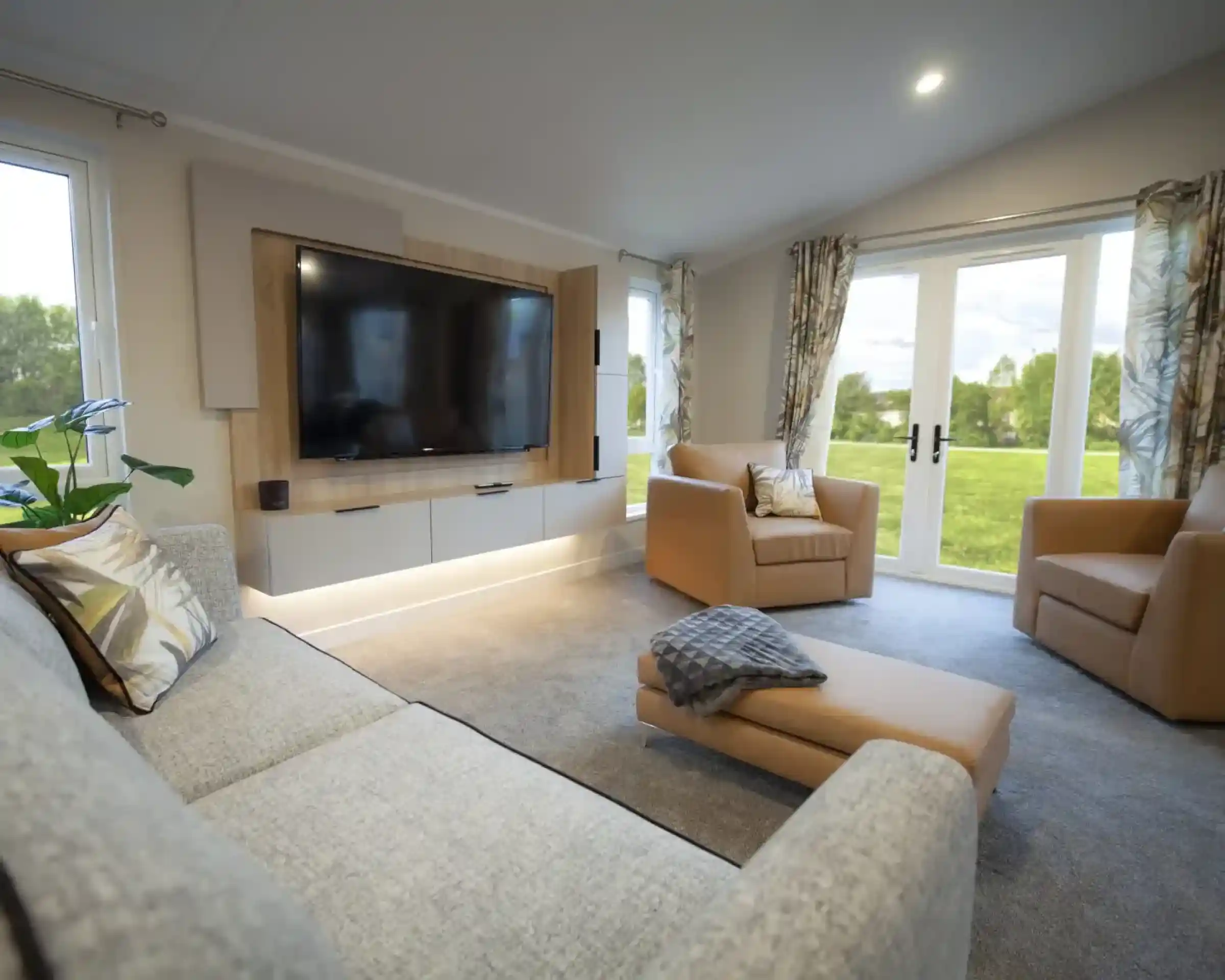 Bright and modern lounge featuring a wall-mounted flat-screen TV with wood panelling, a grey fabric sofa with patterned cushions, two tan armchairs, and a matching ottoman with a throw. Large windows and glass doors with patterned curtains provide views of a green outdoor lawn, filling the room with natural light.