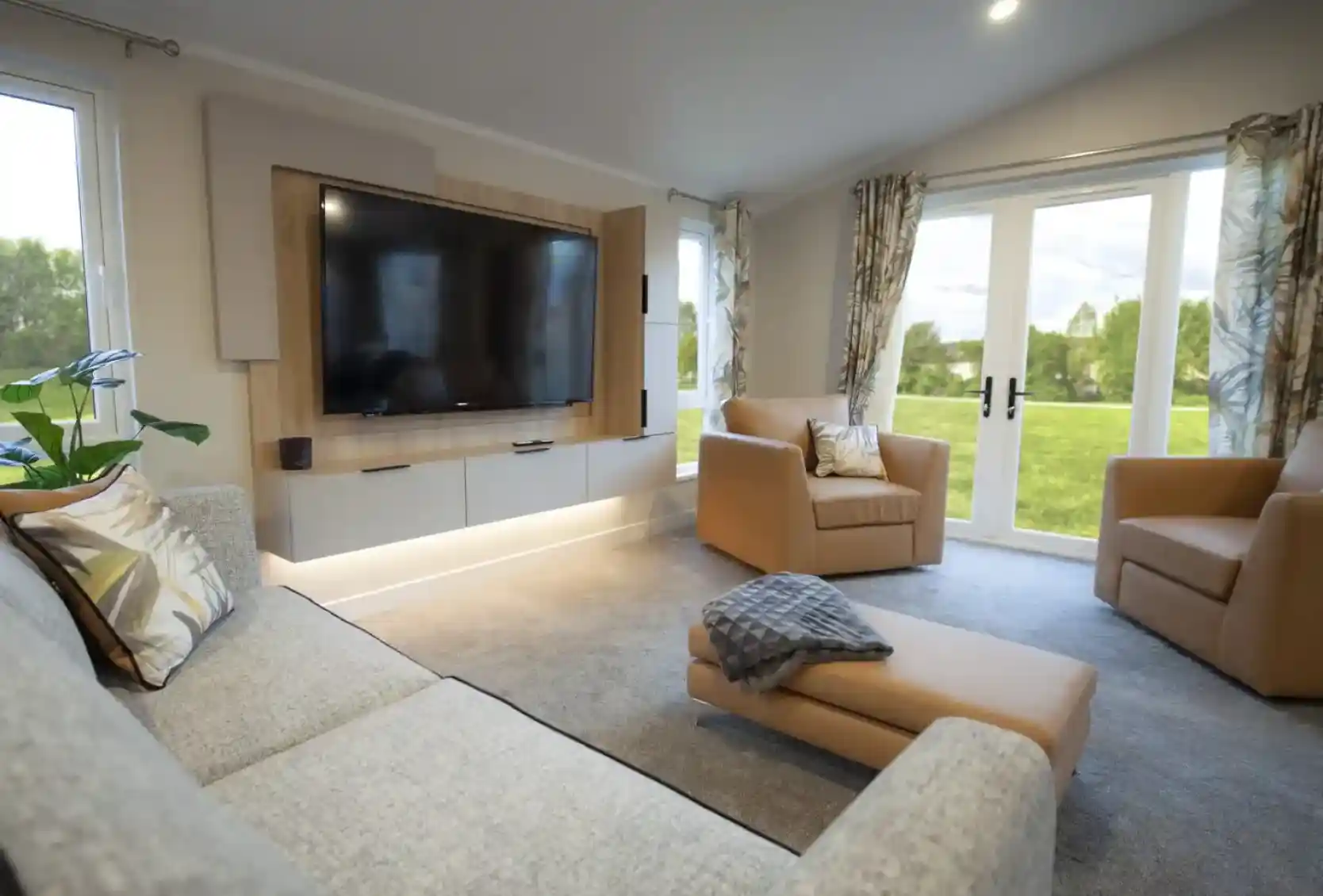 Bright and modern lounge featuring a wall-mounted flat-screen TV with wood panelling, a grey fabric sofa with patterned cushions, two tan armchairs, and a matching ottoman with a throw. Large windows and glass doors with patterned curtains provide views of a green outdoor lawn, filling the room with natural light.