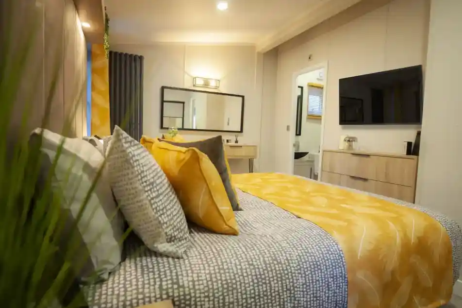 A cosy bedroom featuring a bed with patterned bedding and yellow accent pillows, a dresser with a television, and a doorway leading to a bathroom.