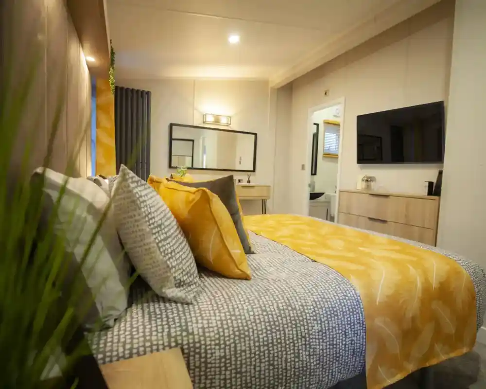A cosy bedroom featuring a bed with patterned bedding and yellow accent pillows, a dresser with a television, and a doorway leading to a bathroom.