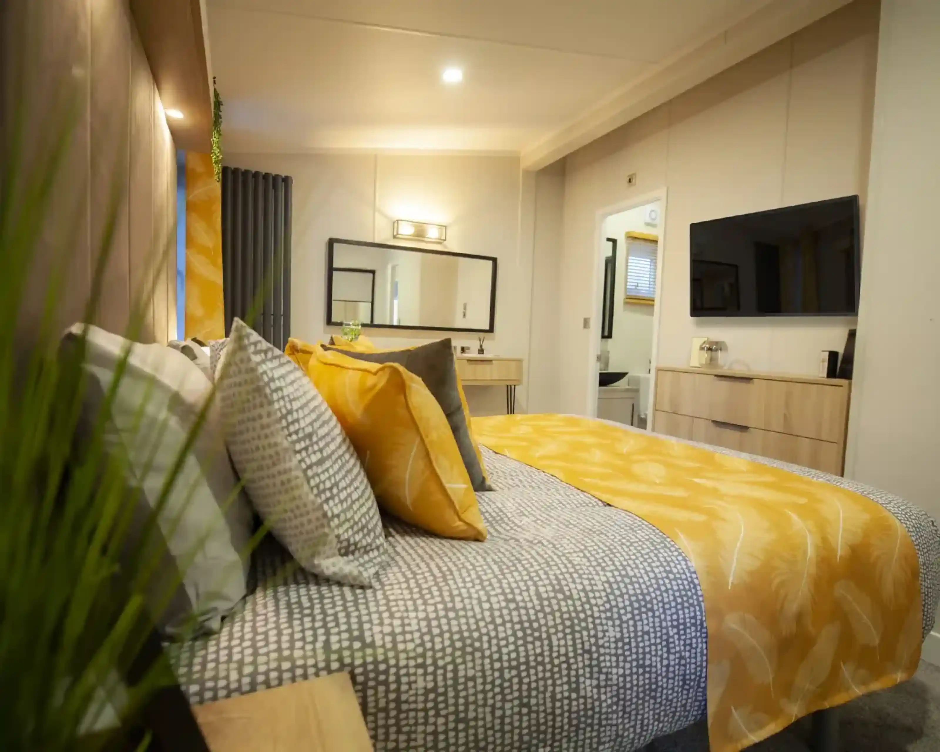 A cosy bedroom featuring a bed with patterned bedding and yellow accent pillows, a dresser with a television, and a doorway leading to a bathroom.