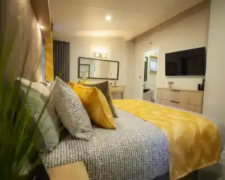 A cosy bedroom featuring a bed with patterned bedding and yellow accent pillows, a dresser with a television, and a doorway leading to a bathroom.