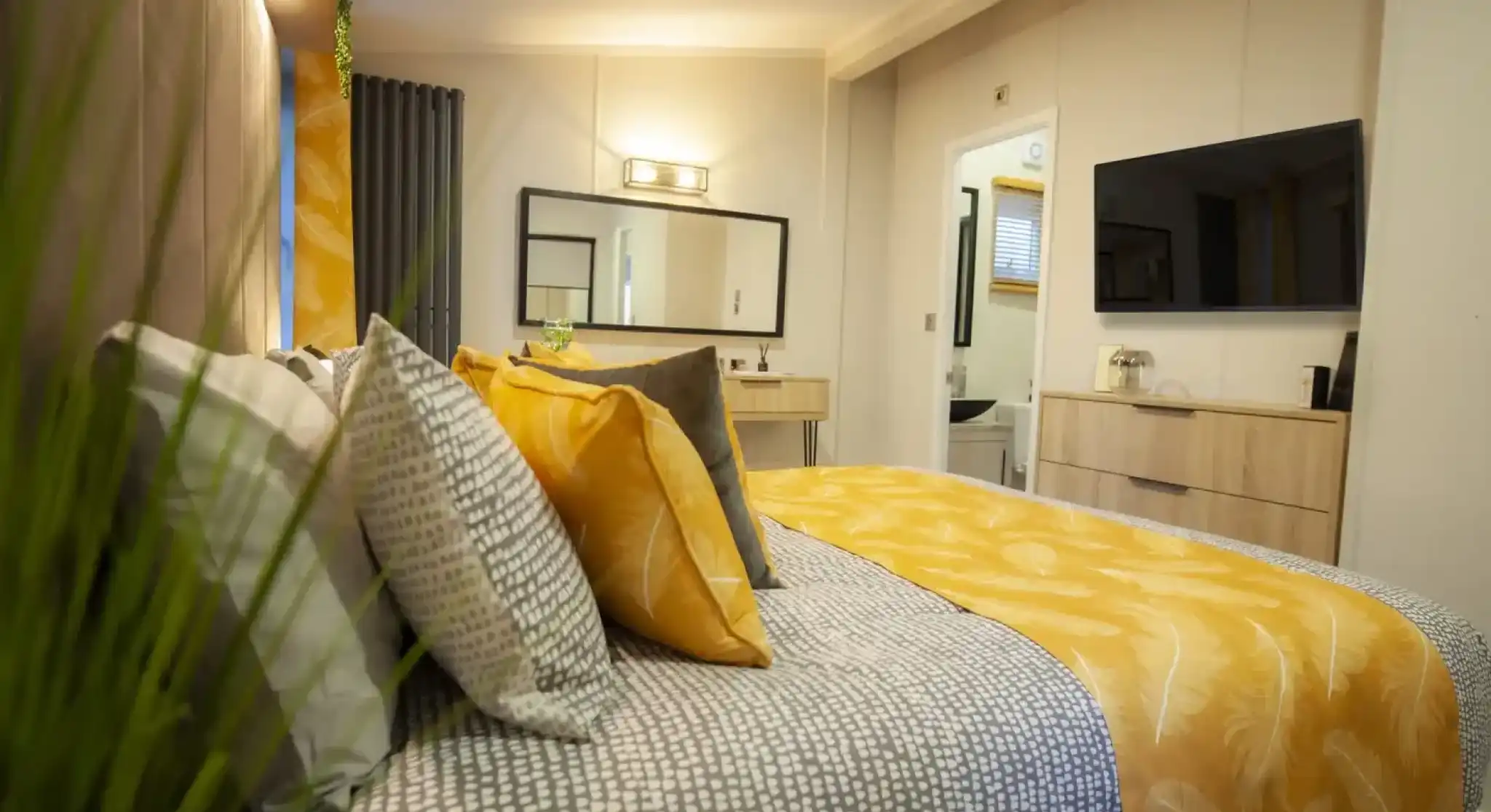 A cosy bedroom featuring a bed with patterned bedding and yellow accent pillows, a dresser with a television, and a doorway leading to a bathroom.