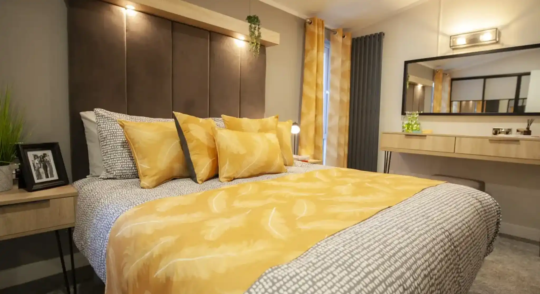 A bedroom with a large bed featuring a yellow feather-patterned duvet and multiple yellow pillows, a dark upholstered headboard with a wooden shelf above, and a vanity with a mirror in the background.