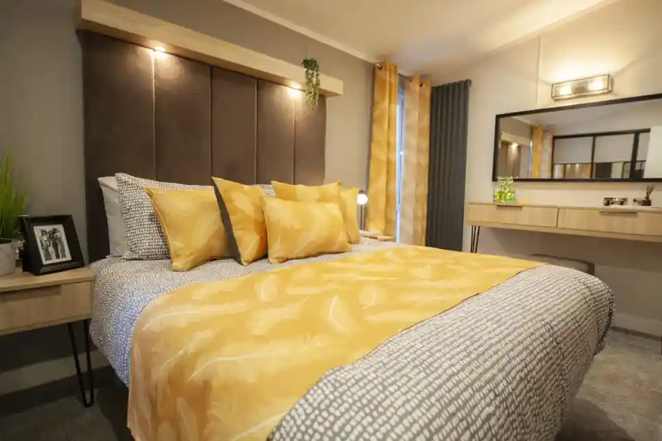 A bedroom with a large bed featuring a yellow feather-patterned duvet and multiple yellow pillows, a dark upholstered headboard with a wooden shelf above, and a vanity with a mirror in the background.