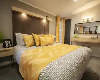 A bedroom with a large bed featuring a yellow feather-patterned duvet and multiple yellow pillows, a dark upholstered headboard with a wooden shelf above, and a vanity with a mirror in the background.
