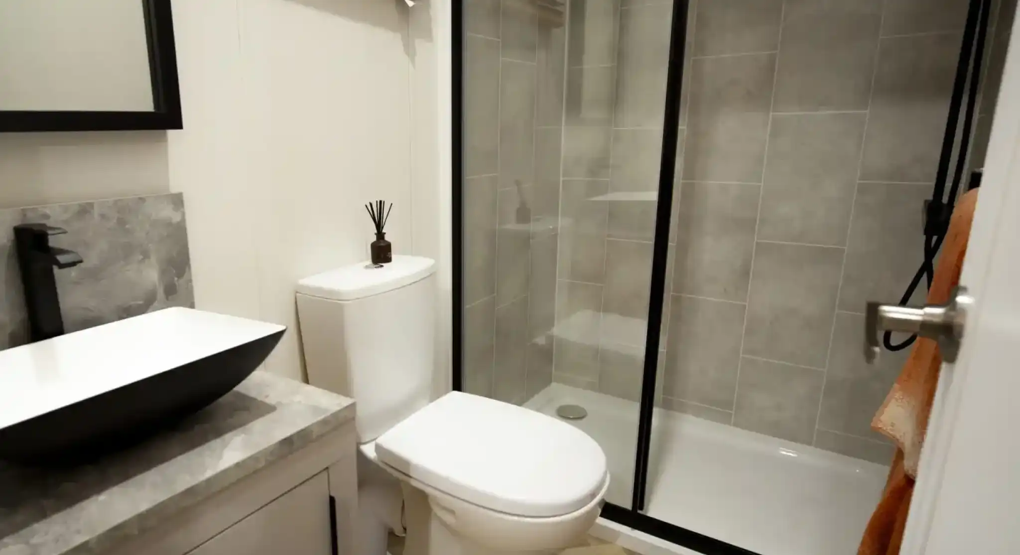 Modern bathroom with a black and white vessel sink, a white toilet, and a glass-enclosed shower with grey tiled walls.