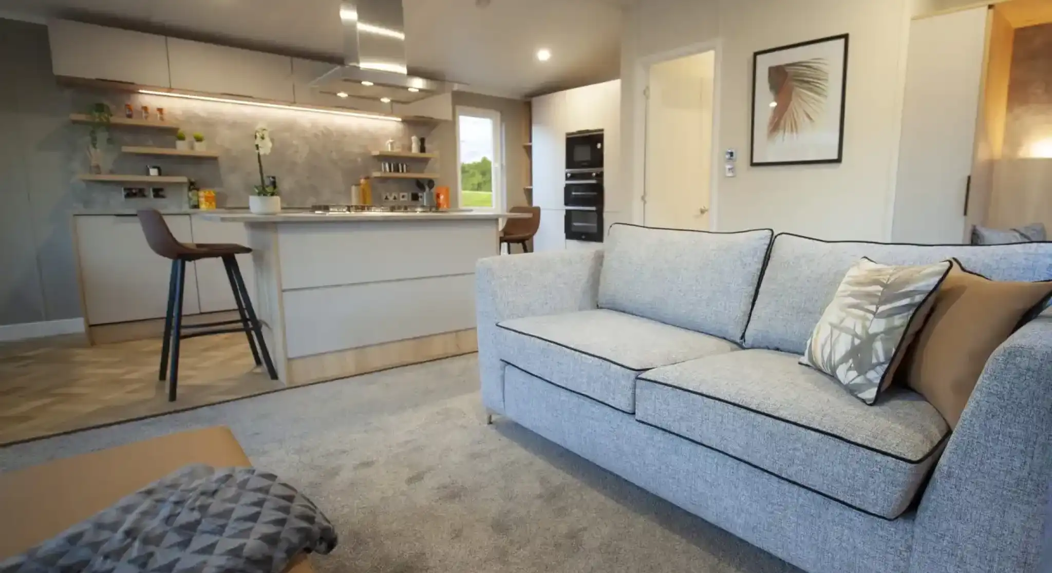 Open-plan living space with a light grey sofa and patterned cushions, a matching ottoman with a throw, and a modern kitchen in the background. The kitchen features a central island with bar stools, overhead extractor fan, built-in ovens, and open shelving with plants and décor. Neutral tones and soft lighting create a contemporary, inviting atmosphere.