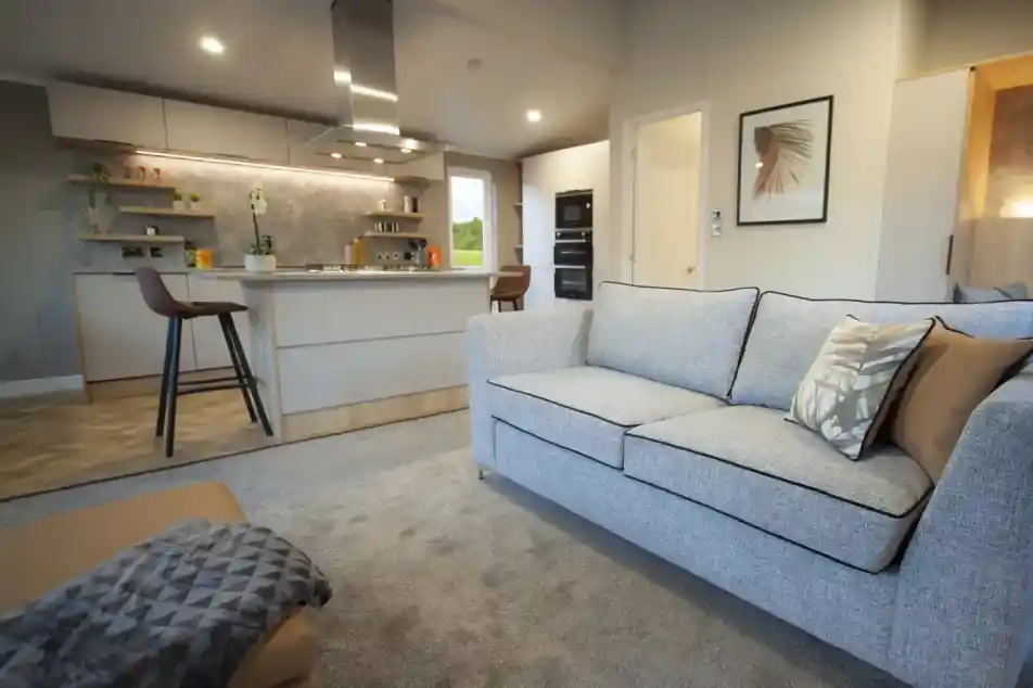 Open-plan living space with a light grey sofa and patterned cushions, a matching ottoman with a throw, and a modern kitchen in the background. The kitchen features a central island with bar stools, overhead extractor fan, built-in ovens, and open shelving with plants and décor. Neutral tones and soft lighting create a contemporary, inviting atmosphere.