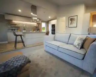 Open-plan living space with a light grey sofa and patterned cushions, a matching ottoman with a throw, and a modern kitchen in the background. The kitchen features a central island with bar stools, overhead extractor fan, built-in ovens, and open shelving with plants and décor. Neutral tones and soft lighting create a contemporary, inviting atmosphere.