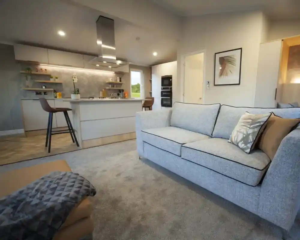 Open-plan living space with a light grey sofa and patterned cushions, a matching ottoman with a throw, and a modern kitchen in the background. The kitchen features a central island with bar stools, overhead extractor fan, built-in ovens, and open shelving with plants and décor. Neutral tones and soft lighting create a contemporary, inviting atmosphere.