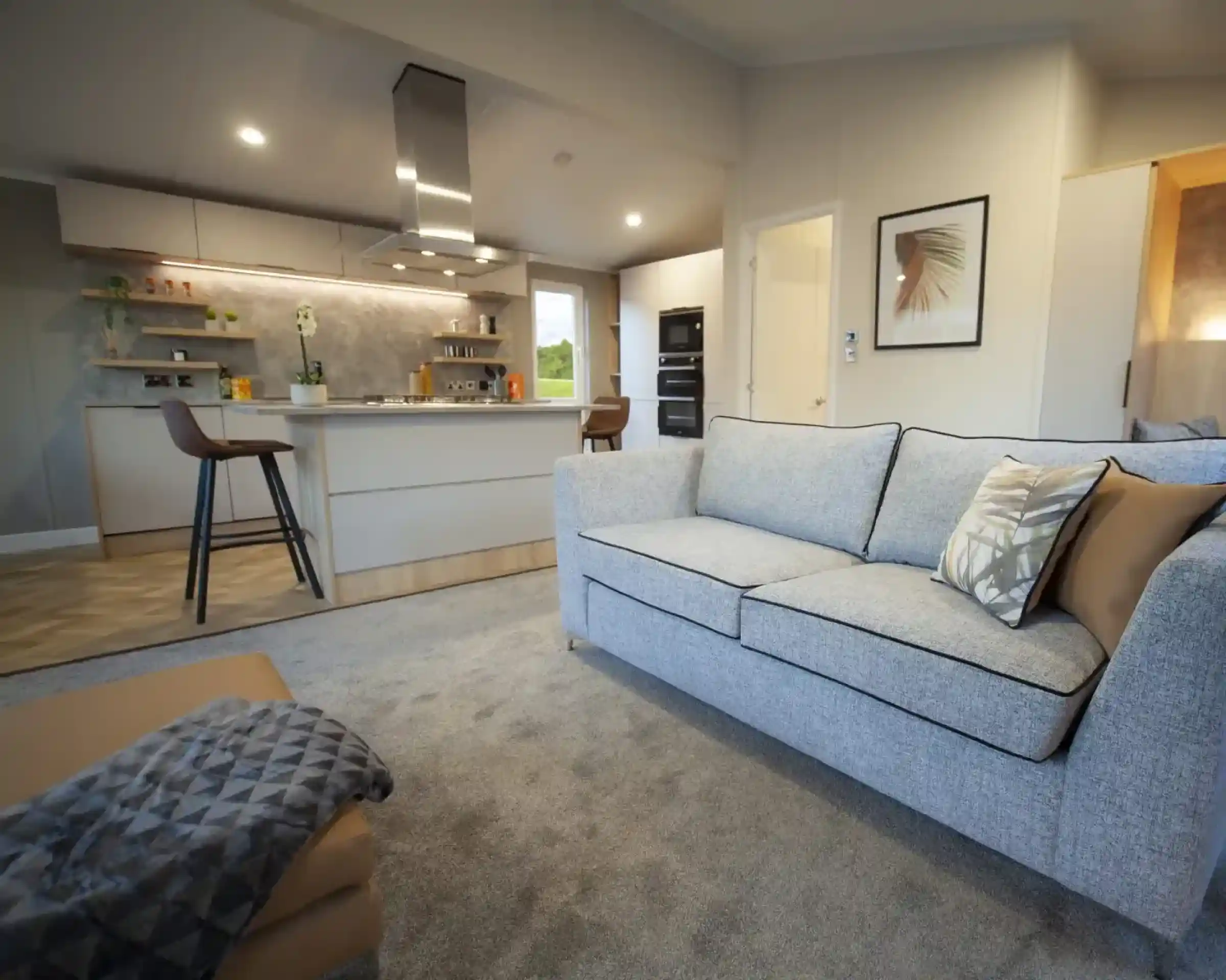 Open-plan living space with a light grey sofa and patterned cushions, a matching ottoman with a throw, and a modern kitchen in the background. The kitchen features a central island with bar stools, overhead extractor fan, built-in ovens, and open shelving with plants and décor. Neutral tones and soft lighting create a contemporary, inviting atmosphere.