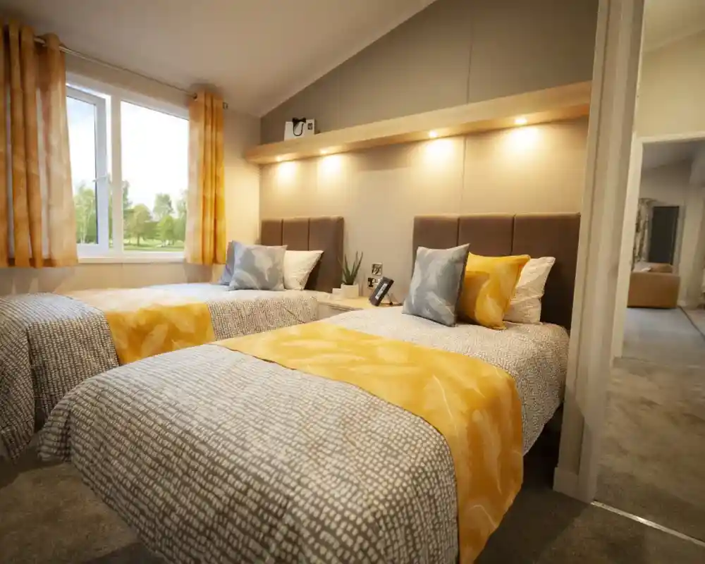 Two twin beds with patterned grey and white bedding are positioned side-by-side in a bedroom with a large window.