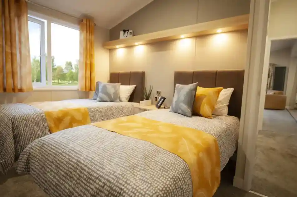 Two twin beds with patterned grey and white bedding are positioned side-by-side in a bedroom with a large window.