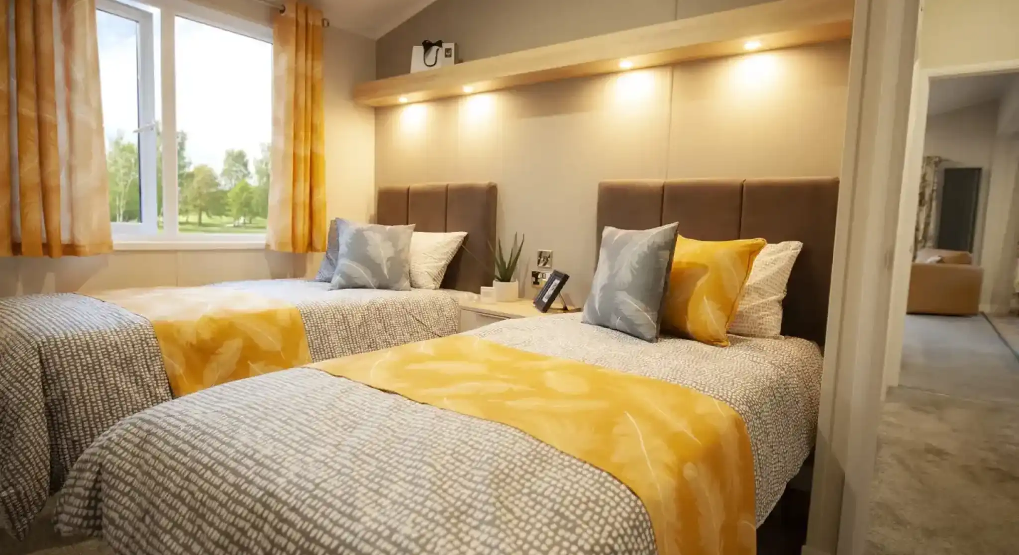 Two twin beds with patterned grey and white bedding are positioned side-by-side in a bedroom with a large window.
