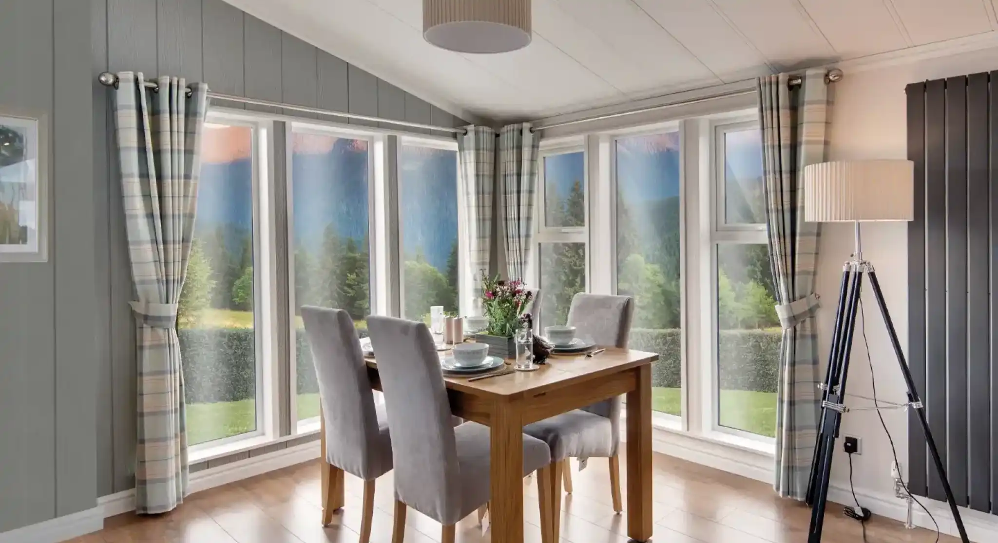 Dining area with wooden table, grey chairs, plaid curtains, and large windows overlooking a scenic landscape.