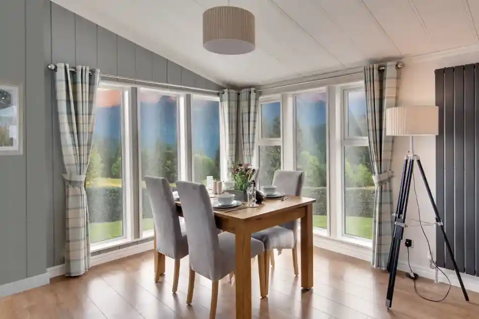 Dining area with wooden table, grey chairs, plaid curtains, and large windows overlooking a scenic landscape.