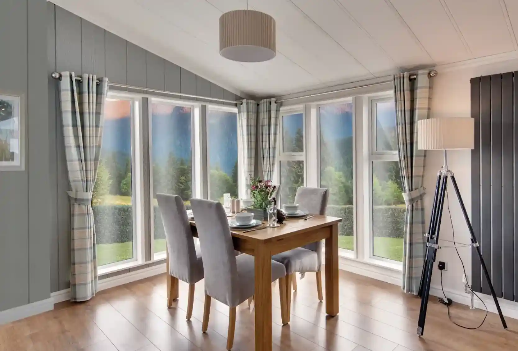 Dining area with wooden table, grey chairs, plaid curtains, and large windows overlooking a scenic landscape.