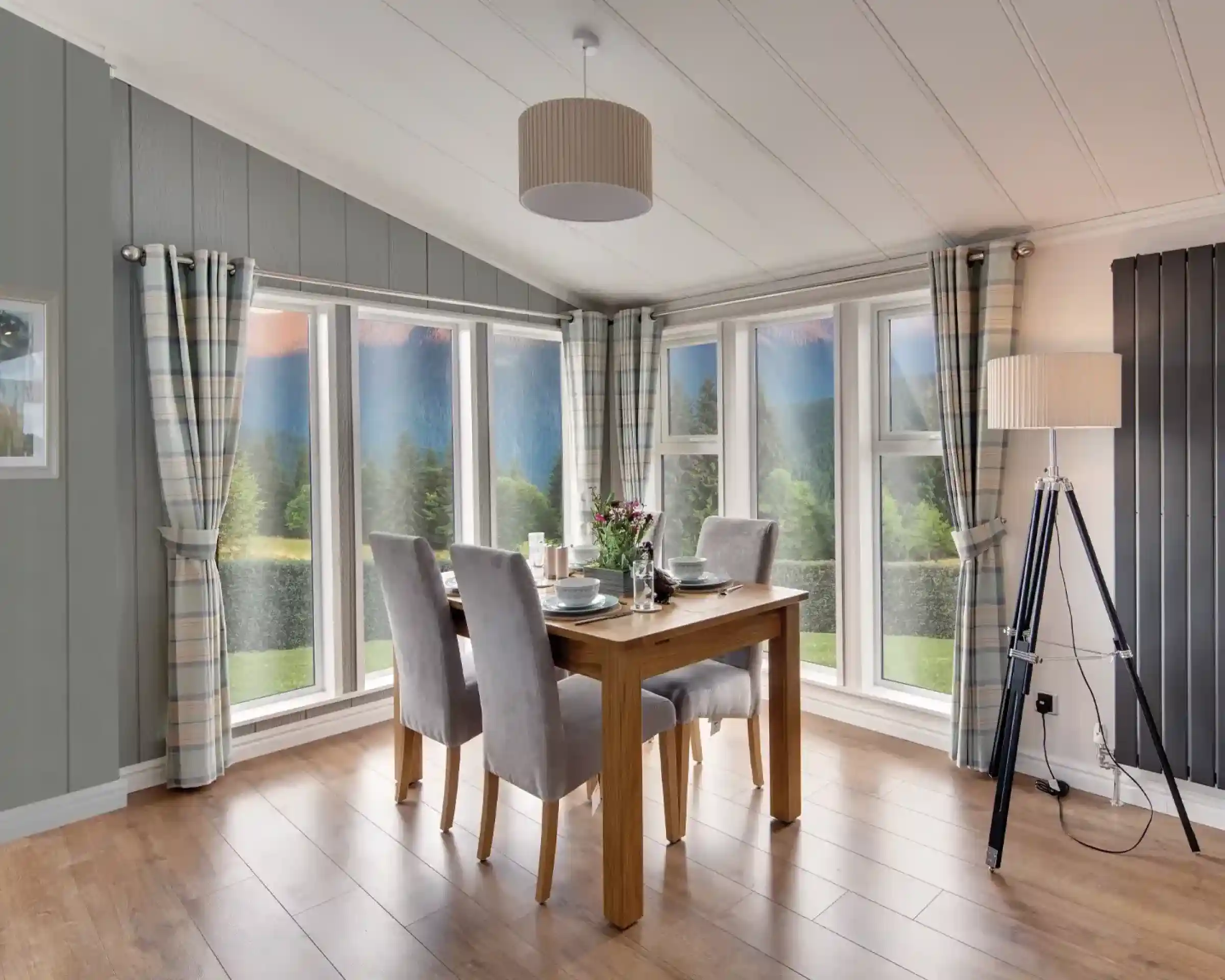 Dining area with wooden table, grey chairs, plaid curtains, and large windows overlooking a scenic landscape.