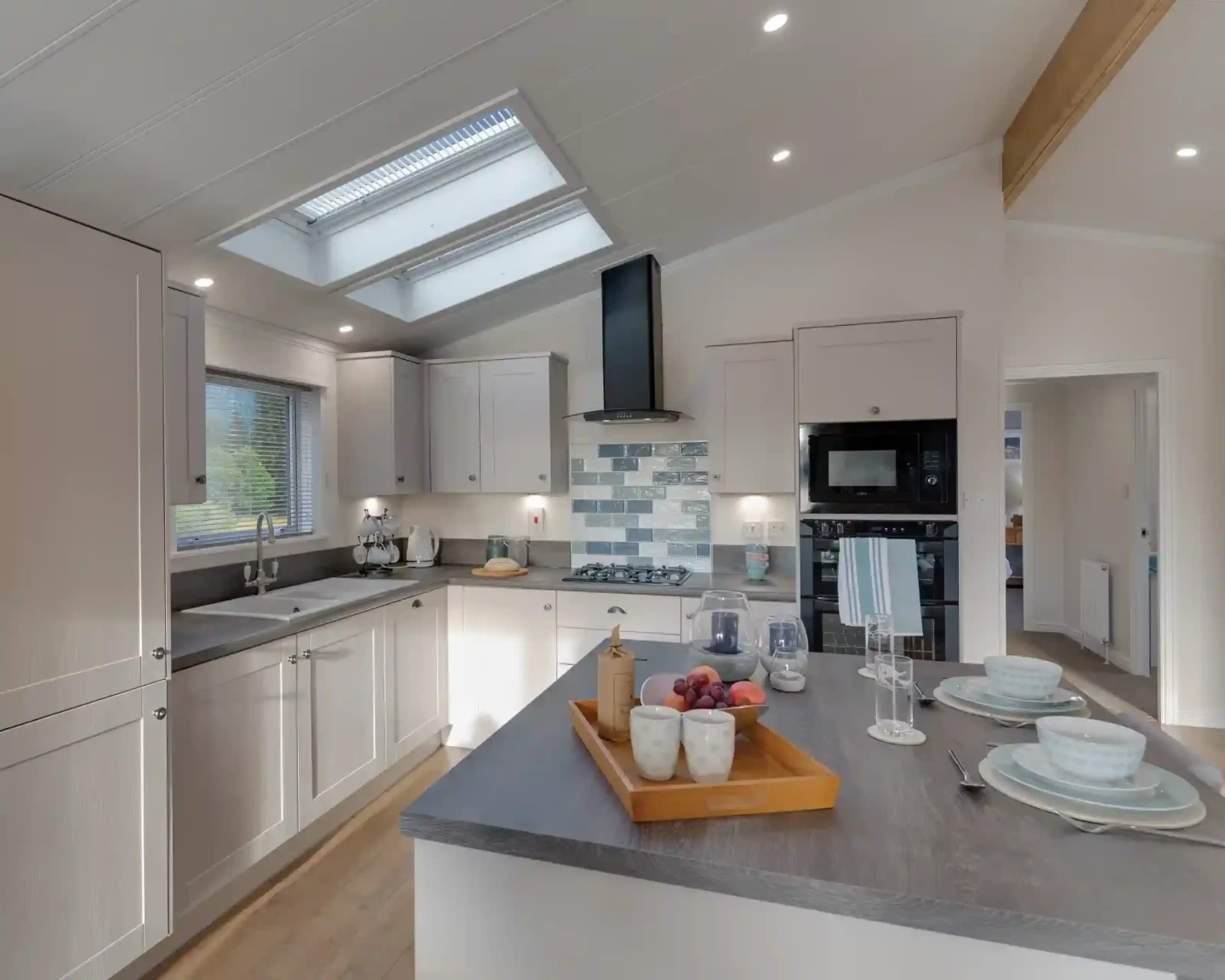 A modern, light-filled kitchen with white cabinetry, dual skylights, a grey island with fruit and place settings, and a view into an adjacent room.