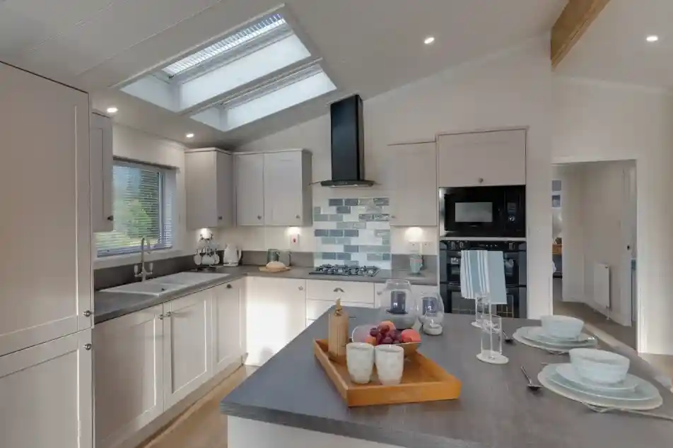 A modern, light-filled kitchen with white cabinetry, dual skylights, a grey island with fruit and place settings, and a view into an adjacent room.