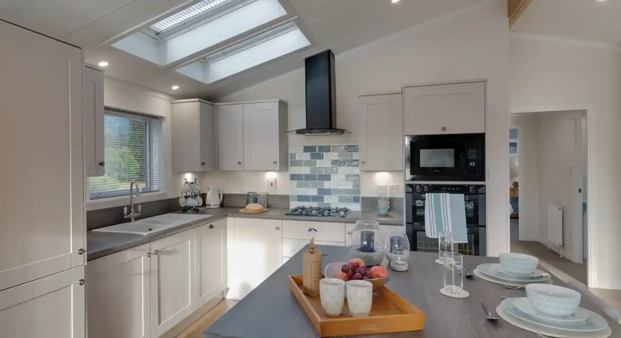 A modern, light-filled kitchen with white cabinetry, dual skylights, a grey island with fruit and place settings, and a view into an adjacent room.