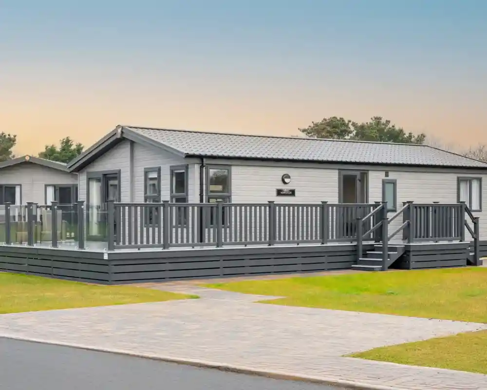 A modern, light grey lodge holiday home with a wrap-around deck and glass railings sits on a grassy lawn with a paved walkway.