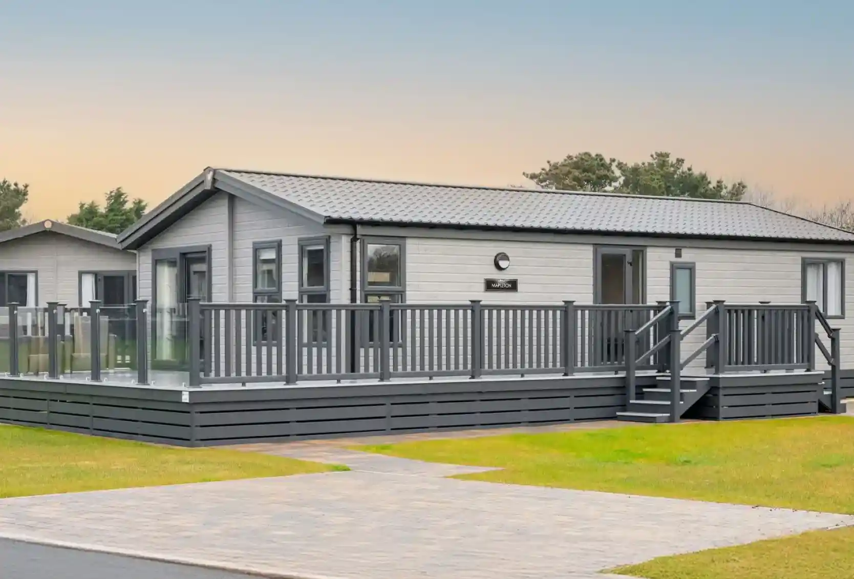 A modern, light grey lodge holiday home with a wrap-around deck and glass railings sits on a grassy lawn with a paved walkway.