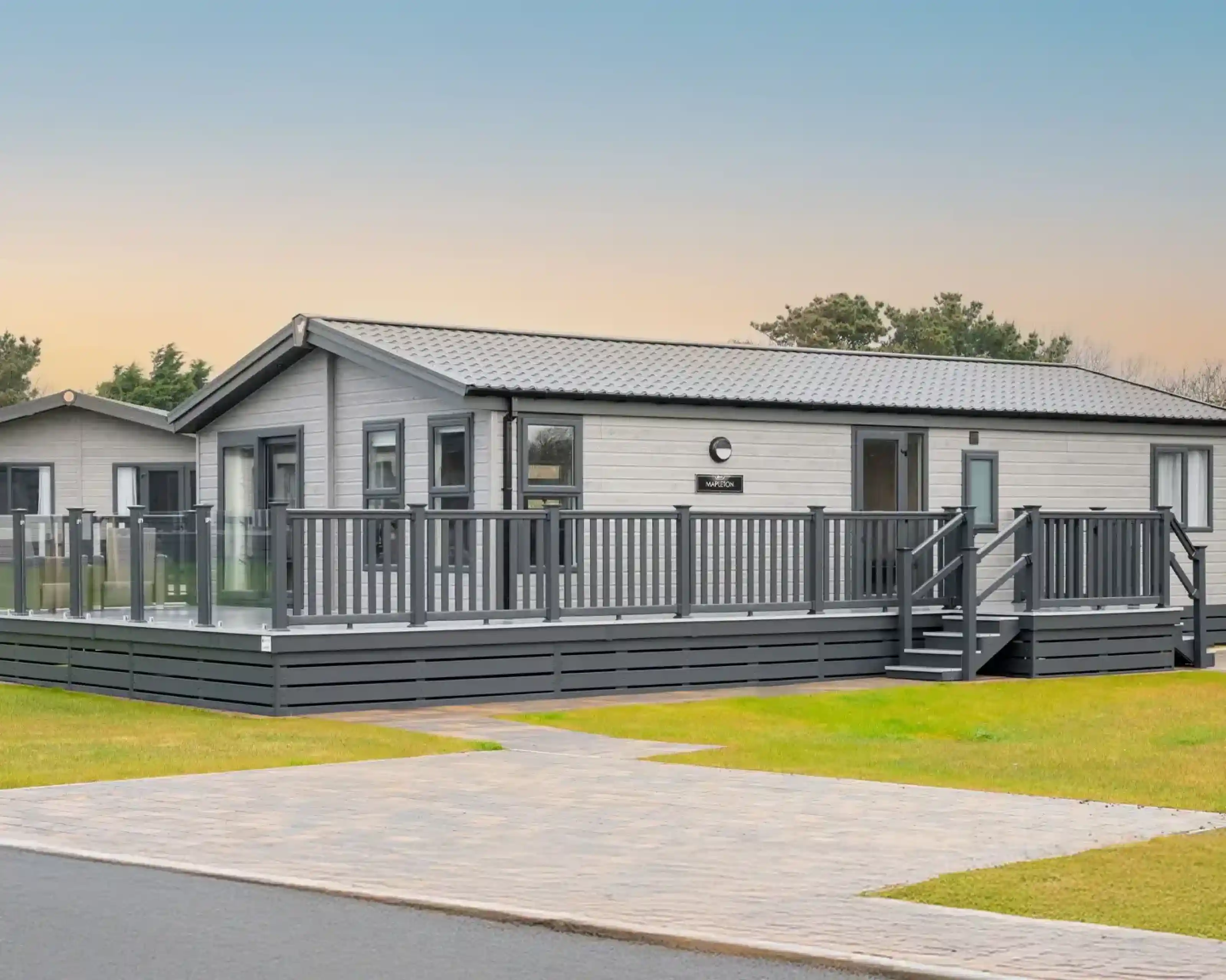 A modern, light grey lodge holiday home with a wrap-around deck and glass railings sits on a grassy lawn with a paved walkway.