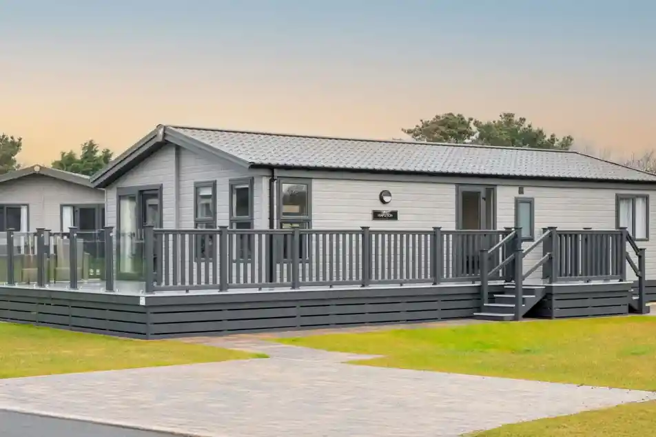 A modern, light grey lodge holiday home with a wrap-around deck and glass railings sits on a grassy lawn with a paved walkway.