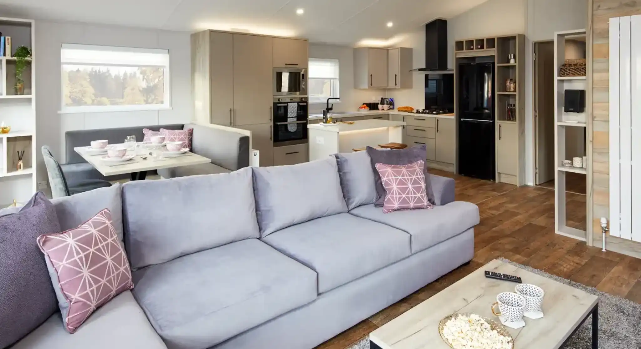 Modern living room with light grey sectional sofa facing a coffee table with popcorn and a modern kitchen with island and dining nook in the background.
