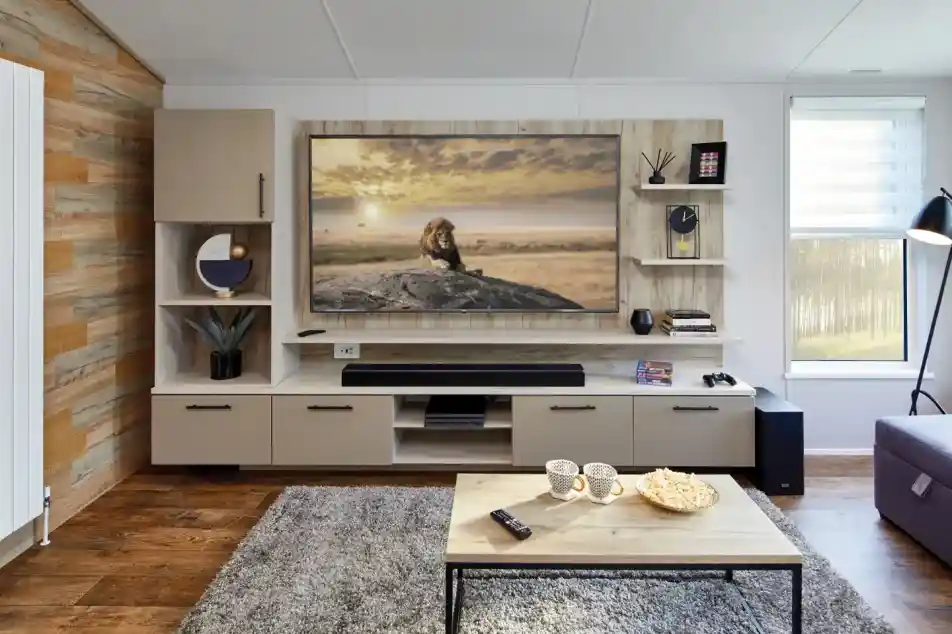 Modern living room with a large TV displaying a lion on a rock, a sound bar, shelving, and a coffee table with popcorn and mugs.