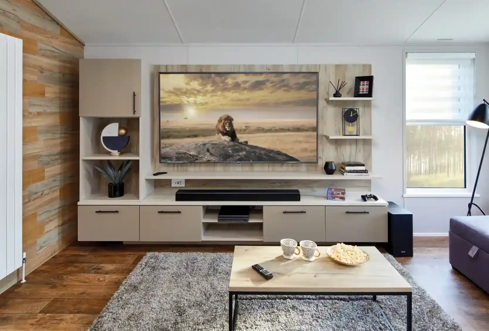 Modern living room with a large TV displaying a lion on a rock, a sound bar, shelving, and a coffee table with popcorn and mugs.
