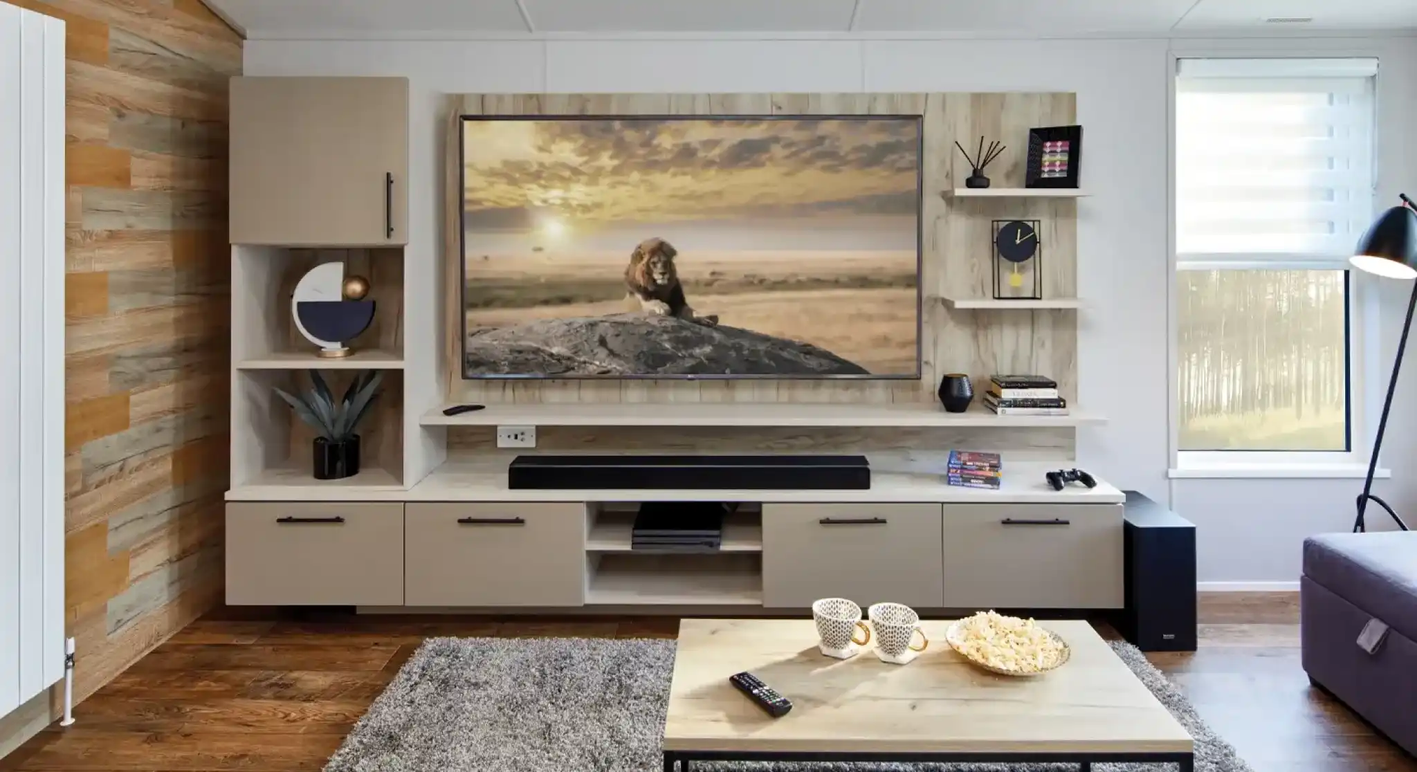 Modern living room with a large TV displaying a lion on a rock, a sound bar, shelving, and a coffee table with popcorn and mugs.