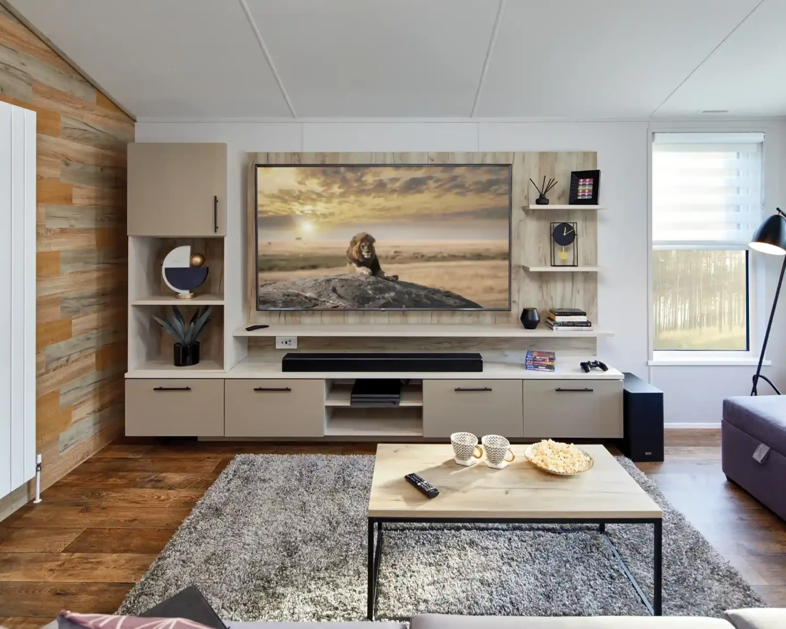 Modern living room with a large TV displaying a lion on a rock, a sound bar, shelving, and a coffee table with popcorn and mugs.