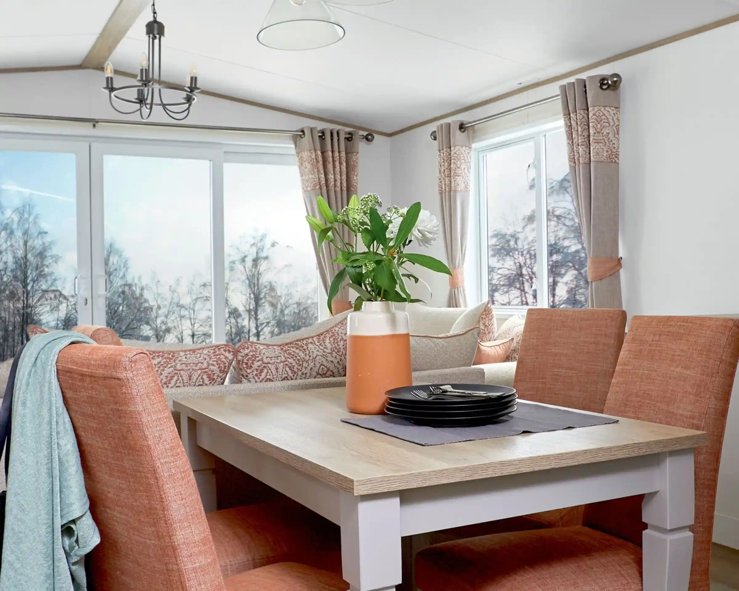 Stylish dining area inside a holiday home or static caravan, featuring a wooden dining table with four upholstered chairs in a warm terracotta color. A decorative vase with flowers and a stack of black plates sit on a table runner. In the background, a cozy L-shaped sofa with patterned cushions and large windows with neutral and coral-toned curtains provide a bright, scenic view of trees outside.