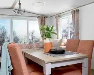 Stylish dining area inside a holiday home or static caravan, featuring a wooden dining table with four upholstered chairs in a warm terracotta color. A decorative vase with flowers and a stack of black plates sit on a table runner. In the background, a cozy L-shaped sofa with patterned cushions and large windows with neutral and coral-toned curtains provide a bright, scenic view of trees outside.