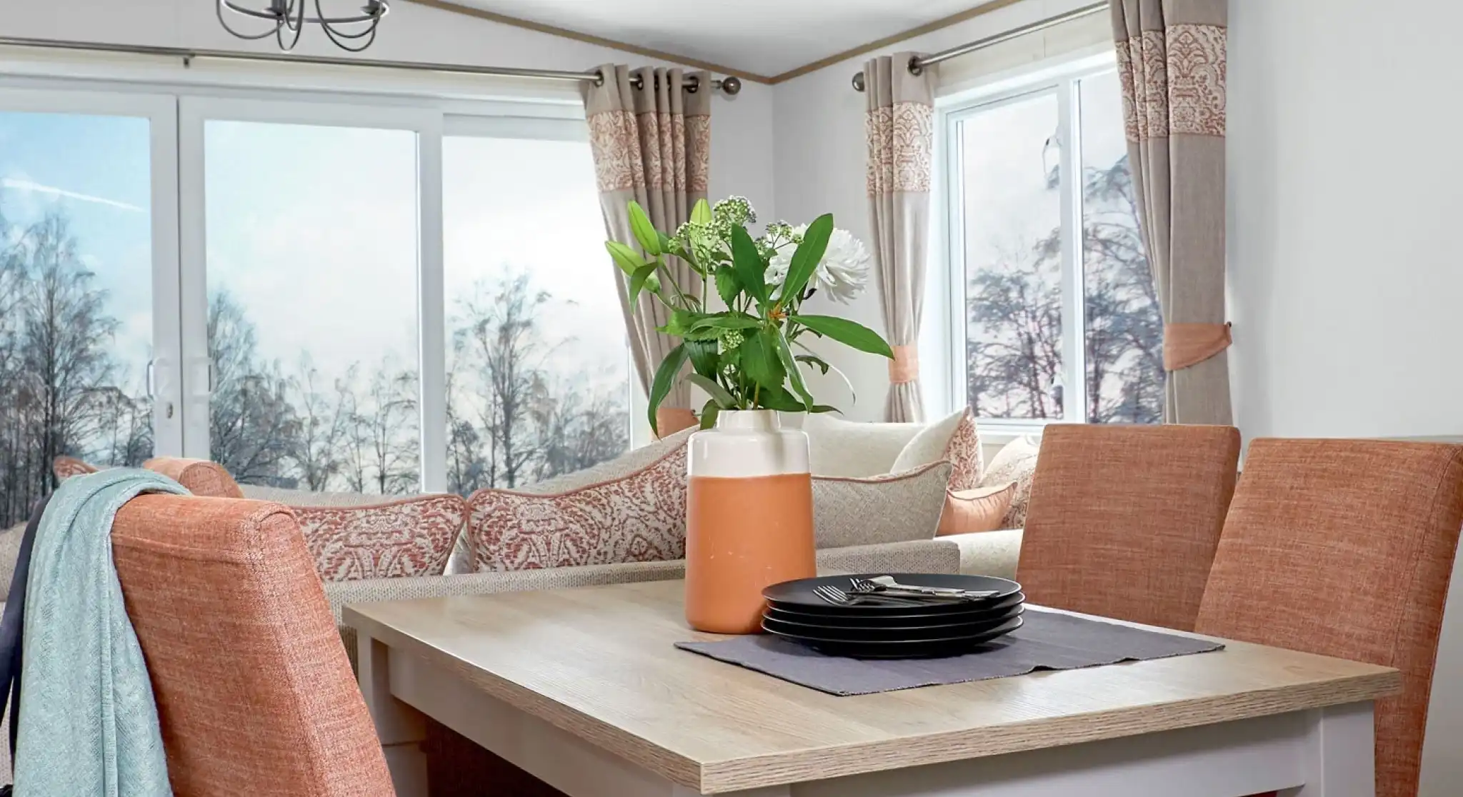 Stylish dining area inside a holiday home or static caravan, featuring a wooden dining table with four upholstered chairs in a warm terracotta color. A decorative vase with flowers and a stack of black plates sit on a table runner. In the background, a cozy L-shaped sofa with patterned cushions and large windows with neutral and coral-toned curtains provide a bright, scenic view of trees outside.