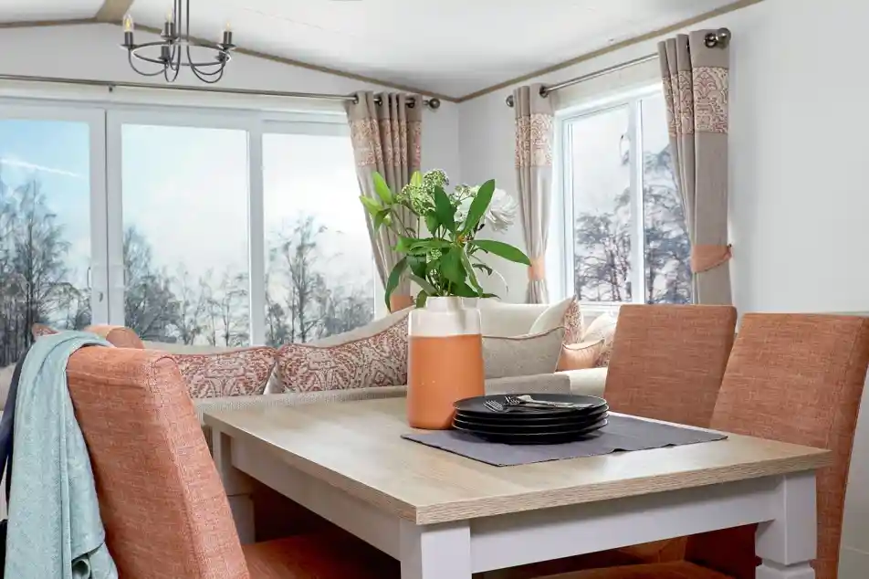 Stylish dining area inside a holiday home or static caravan, featuring a wooden dining table with four upholstered chairs in a warm terracotta color. A decorative vase with flowers and a stack of black plates sit on a table runner. In the background, a cozy L-shaped sofa with patterned cushions and large windows with neutral and coral-toned curtains provide a bright, scenic view of trees outside.