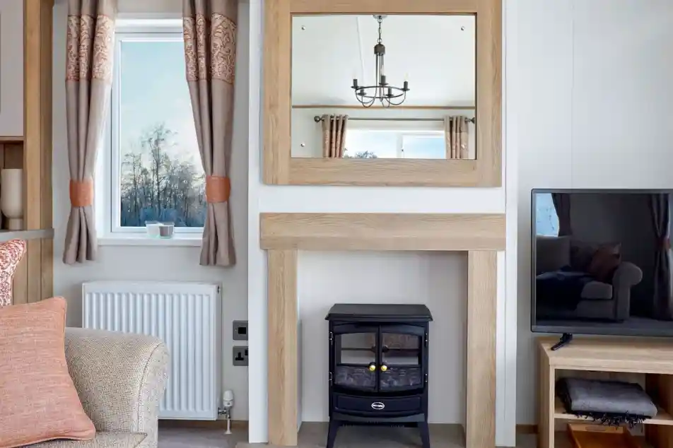 Stylish lounge area in the ABI Ambleside holiday home, featuring a wood-framed mirror above a modern fireplace with an electric log-effect stove, flanked by a window with patterned curtains and a flat-screen TV on a wooden media unit.