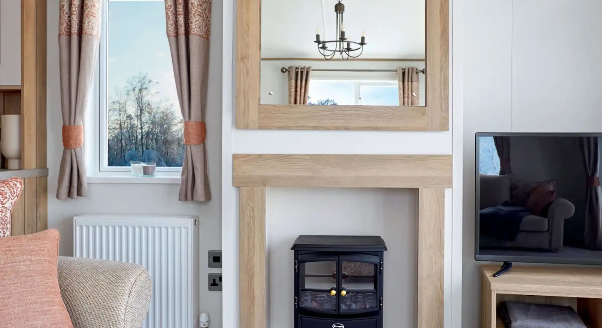 Stylish lounge area in the ABI Ambleside holiday home, featuring a wood-framed mirror above a modern fireplace with an electric log-effect stove, flanked by a window with patterned curtains and a flat-screen TV on a wooden media unit.