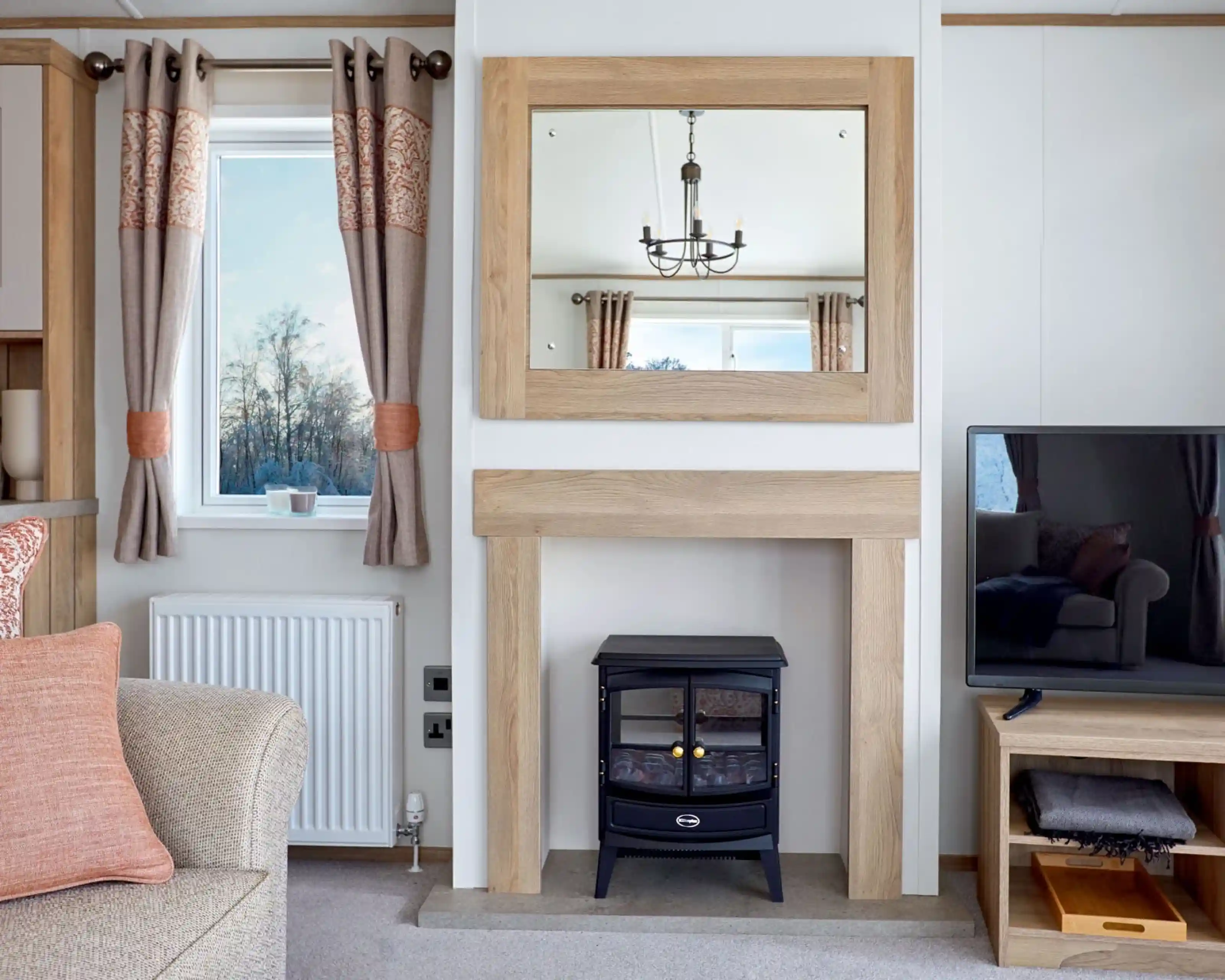 Stylish lounge area in the ABI Ambleside holiday home, featuring a wood-framed mirror above a modern fireplace with an electric log-effect stove, flanked by a window with patterned curtains and a flat-screen TV on a wooden media unit.